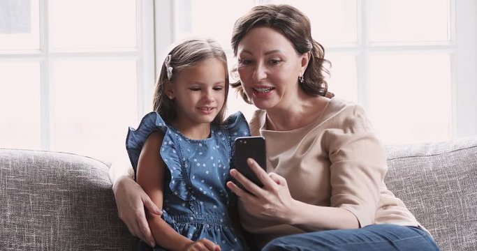 Mature grandma hug child granddaughter using smartphone at home