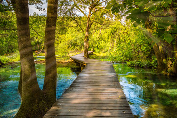 Fototapeta na wymiar Wooden footpath over river in forest of Krka National Park, Croatia. Beautiful scene with trees, water and sunrays.
