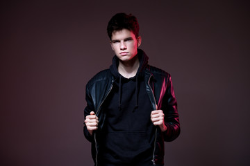 Attractive young guy posing in studio. Young caucasian guy model in black clothes posing in the studio on a dark burgundy background with red backlight. Man in a leather jacket