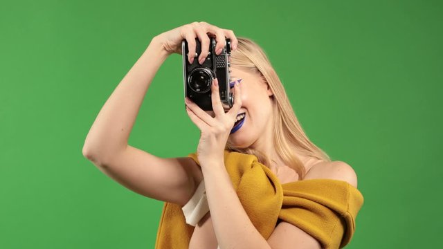 Young blonde woman with a camera in her hands makes a photo and smiles at the camera on an isolated green background, concept of a traveler