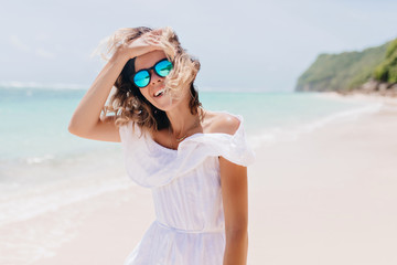 Fototapeta na wymiar Gorgeous girl in white dress touching her hair on sea background. Tanned carefree woman in sunglasses expressing happiness in her vacation.