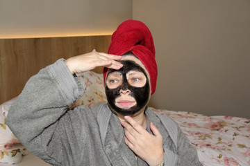 a woman with a black cosmetic mask in a towel on her head sits on the bed in a bathrobe and touches...