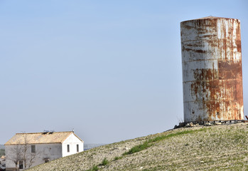 Old and rusty cylindrical water tank on a hill next to a house in the countryside