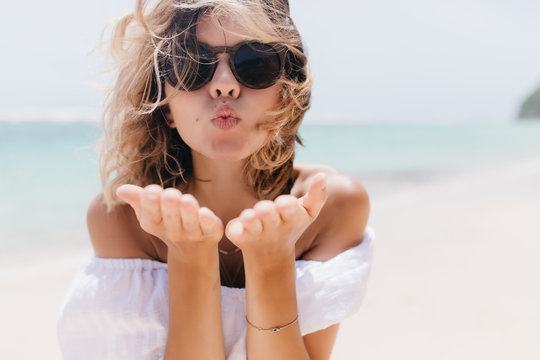 Portrait of good-humoured female model in dark sunglasses standing at sea coast. Outdoor photo of romantic lady posing near ocean with kissing face expression.