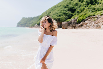 Fototapeta na wymiar Beautiful young woman in white attire standing on tropical background. Outdoor shot of winsome tanned girl in dress posing at sandy beach.