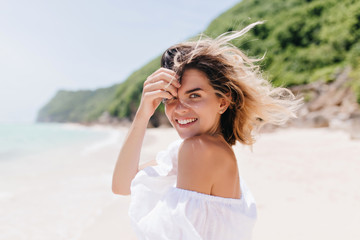 Dreamy tanned woman looking over shoulder while standing at sea coast. Adorable blonde girl posing with pleasure at tropical island in hot day.