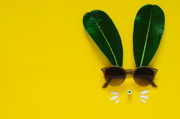 Rabbit face made of green leaves and flowers with sunglasses on yellow background. Flat lay minimal concept for Easter.