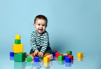 Little baby boy in stylish casual clothing barefoot sitting on floor, playing with colorful toy cubes, showing tongue