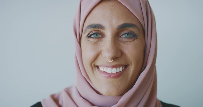 Close up portrait of a young progressive muslim woman wearing a nose ring and a pink traditional hijab head scarf, smiling into the camera laughing with green eyes