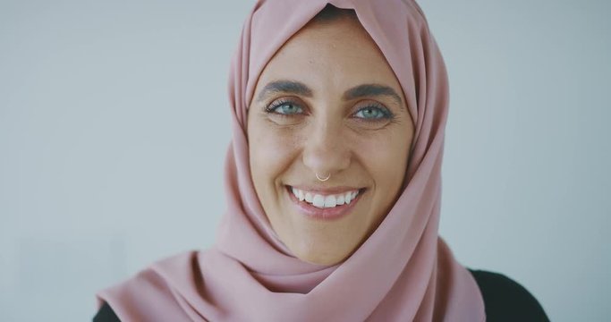 Close up portrait of a young progressive muslim woman wearing a nose ring and a pink traditional hijab head scarf, smiling into the camera laughing with green eyes