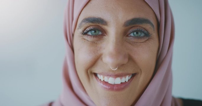 Close up portrait of a young progressive muslim woman wearing a nose ring and a pink traditional hijab head scarf, smiling into the camera laughing with green eyes