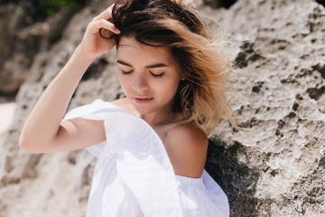 Naklejka premium Relaxed girl in vintage white dress standing on mountain background. Ecstatic dark-haired woman with tanned skin posing near rock.