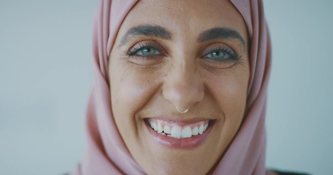 Close up portrait of a young progressive muslim woman wearing a nose ring and a pink traditional hijab head scarf, smiling into the camera laughing with green eyes