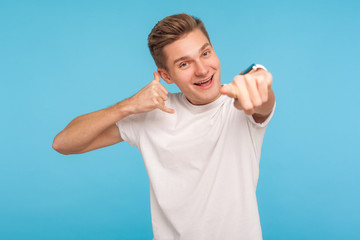 Hey you, call me! Portrait of handsome positive young man in white t-shirt making telephone gesture and pointing to camera, waiting for communication. indoor studio shot isolated on blue background