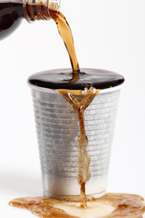 Pouring cola from bottle into overflowing plastic cup. Isolated on white background.