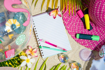 Blank writing note pad with pink and green pen on sand, surrounded by green palm leafs, sea shelles, pink hat, inflatable toys. Summer beach background, flat lay