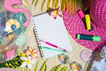 Obraz premium Blank writing note pad with pink and green pen on sand, surrounded by green palm leafs, sea shelles, pink hat, inflatable toys. Summer beach background, flat lay