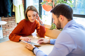 portrait of beautiful young caucasian couple sitting in modern cafe using tablet. handsome man in shirt and attratctive lady in blouse have lunch together and watch something interesting on tablet