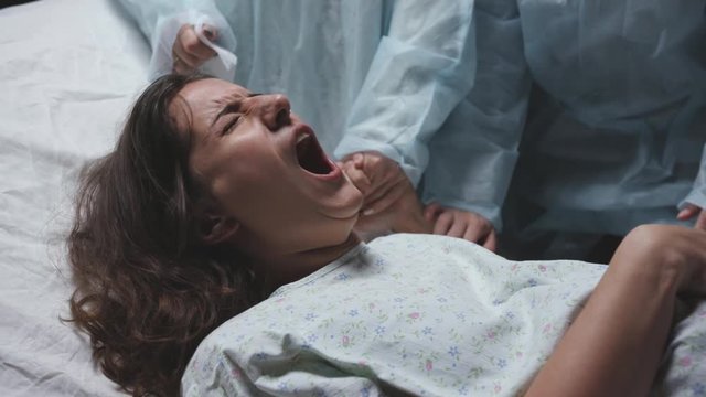 young mother screams and cries during childbirth, maternity hospital, strong pain, caucasian woman contractions, obstetricians gynecologists and midwives support