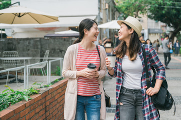 two smiling asian female teenagers walking and holding disposable cups by red brick garden beside. group of happy korean women friends relax in village with paper cup of coffee chatting and laughing
