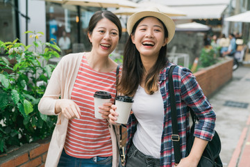 Group of young women tourists stroll through streets of old town. girl friends travelers holding paper cups with coffee in hands on warm summer day. Good mood two ladies laughing looking aside.