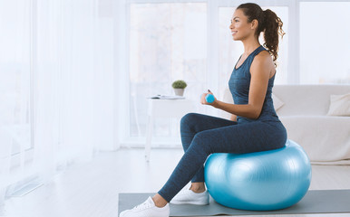 Attractive Hispanic lady with dumbbells sitting on fitness ball at home, copy space