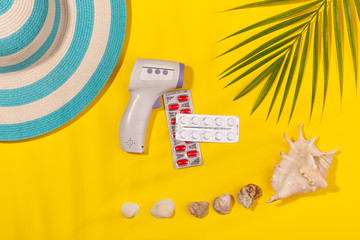 Top view on infrared thermometer, white and red pills in blister pack, blue hat, seashells, palm...