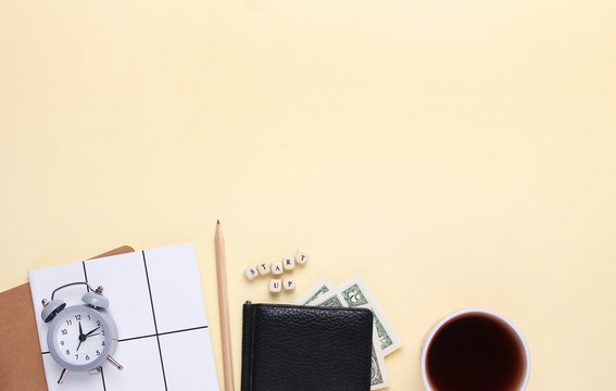Creative flat lay business concept. Notebook with a pencil, wallet,alarm clock,cup of coffee on a beige background with the word start up of wooden letters. Top view, minimalism
