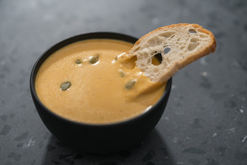 Pumpkin cream soup with pumpkin seeds and baguette crouton in a black bowl on concrete background