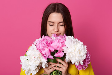 Indoor shot of beautiful brunette girl holding big bouquet with white and rose peonies, attractive woman smells flowers in herhands, lady dresses yellow dress, posing against pink background.