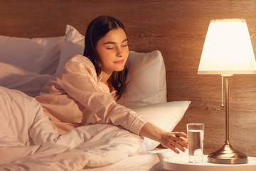 Beautiful young woman taking glass of water from bedside table in evening