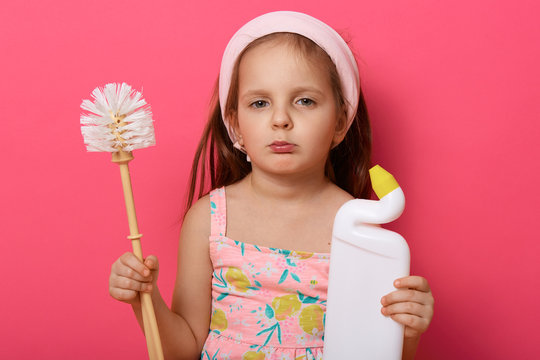 Pretty little girl wearing summer dress wants to halp her mommy to wash toilet, holding brush and detergent, looks at camera with pouty lips, being forbidded to do cleaning up, poses against rosy wall
