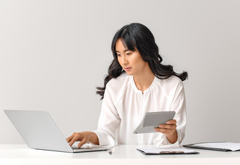 Beautiful Asian secretary at table against light background
