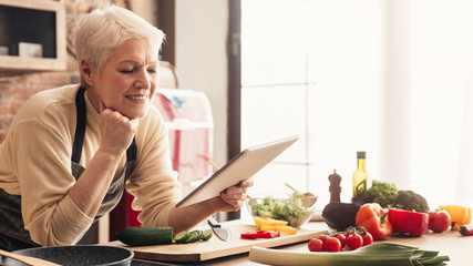 Cheerful senior woman using digital tablet at kitchen, reading news