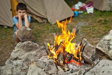 The boy sits by the fire. Sharp on high heat. Baby on tent background.