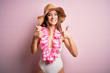 Young beautiful brunette woman on vacation wearing swimsuit and Hawaiian flowers lei success sign doing positive gesture with hand, thumbs up smiling and happy. Cheerful expression and winner gesture.
