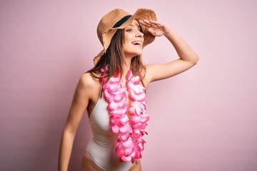 Young beautiful brunette woman on vacation wearing swimsuit and Hawaiian flowers lei very happy and smiling looking far away with hand over head. Searching concept.