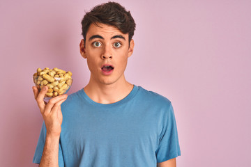 Teenager boy holding a bunch of peanunts over isolated pink background scared in shock with a surprise face, afraid and excited with fear expression