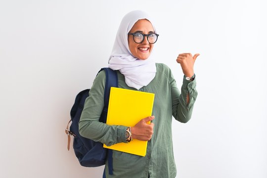 Young Arab student woman wearing hijab and backpack holding a book over isolated background pointing and showing with thumb up to the side with happy face smiling