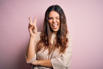 Young beautiful brunette woman wearing casual sweater standing over pink background smiling with happy face winking at the camera doing victory sign with fingers. Number two.