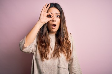 Young beautiful brunette woman wearing casual sweater standing over pink background doing ok gesture shocked with surprised face, eye looking through fingers. Unbelieving expression.