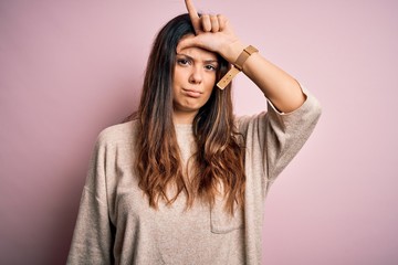 Obraz na płótnie Canvas Young beautiful brunette woman wearing casual sweater standing over pink background making fun of people with fingers on forehead doing loser gesture mocking and insulting.