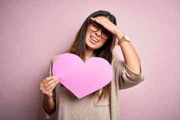 Young beautiful brunette romantic woman holding big heart paper celebrating valentine day stressed...