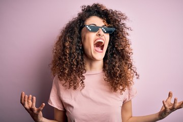 Young beautiful woman with curly hair and piercing wearing funny thug life sunglasses crazy and mad shouting and yelling with aggressive expression and arms raised. Frustration concept.