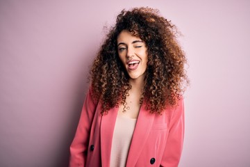 Young beautiful businesswoman with curly hair and piercing wearing elegant jacket winking looking...