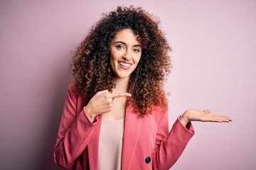 Young beautiful businesswoman with curly hair and piercing wearing elegant jacket amazed and smiling to the camera while presenting with hand and pointing with finger.
