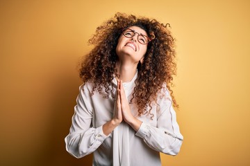 Young beautiful brunette woman with curly hair and piercing wearing shirt and glasses begging and praying with hands together with hope expression on face very emotional and worried. Begging.