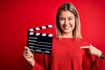 Young beautiful woman holding clapboard standing over isolated red background with surprise face...