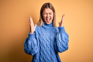 Young beautiful blonde woman wearing turtleneck sweater over yellow isolated background celebrating mad and crazy for success with arms raised and closed eyes screaming excited. Winner concept