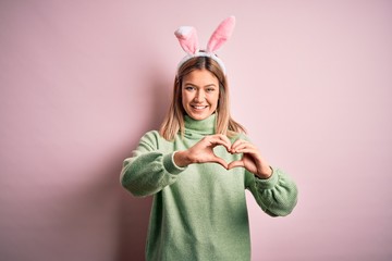 Young beautiful woman wearing easter rabbit ears standing over isolated pink background smiling in love doing heart symbol shape with hands. Romantic concept.